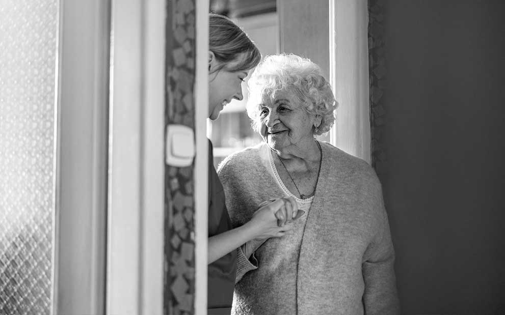 Pflegerin hält lächelnd Hand von Seniorin in Türrahmen